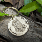Gold keychain with owl engraving on wood, surrounded by green leaves. Customizable text space available.