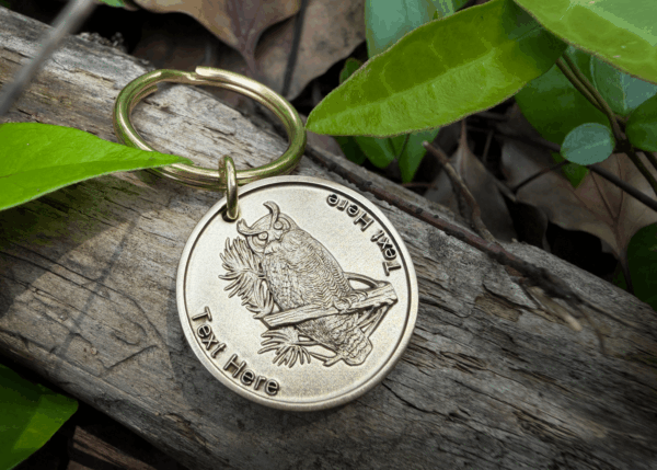 Gold keychain with owl engraving on wood, surrounded by green leaves. Customizable text space available.