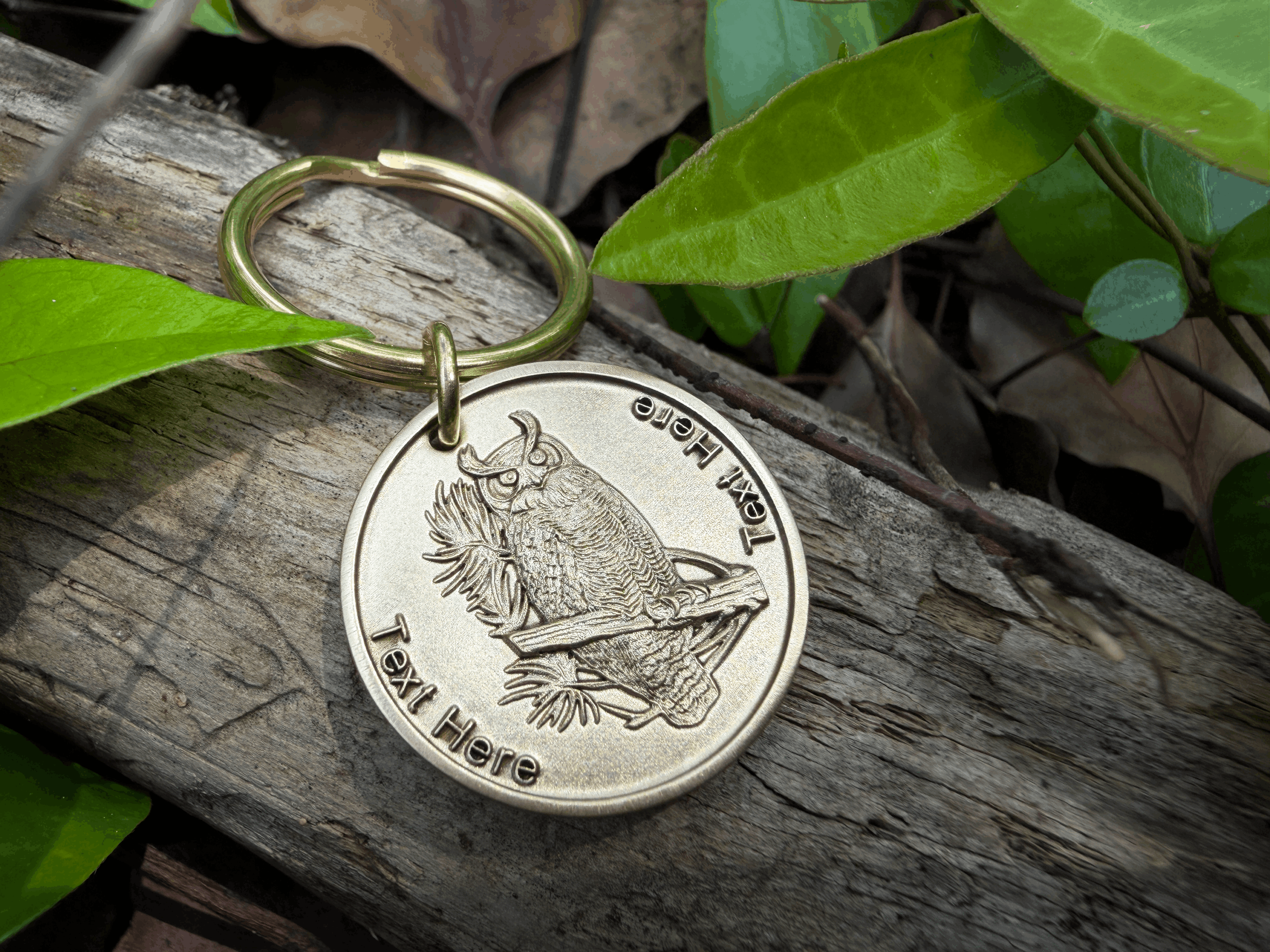 Gold keychain with owl engraving on wood, surrounded by green leaves. Customizable text space available.