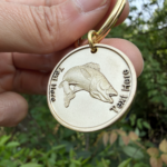 Gold keychain with engraved fish and customizable Text Here displayed against a leafy background.