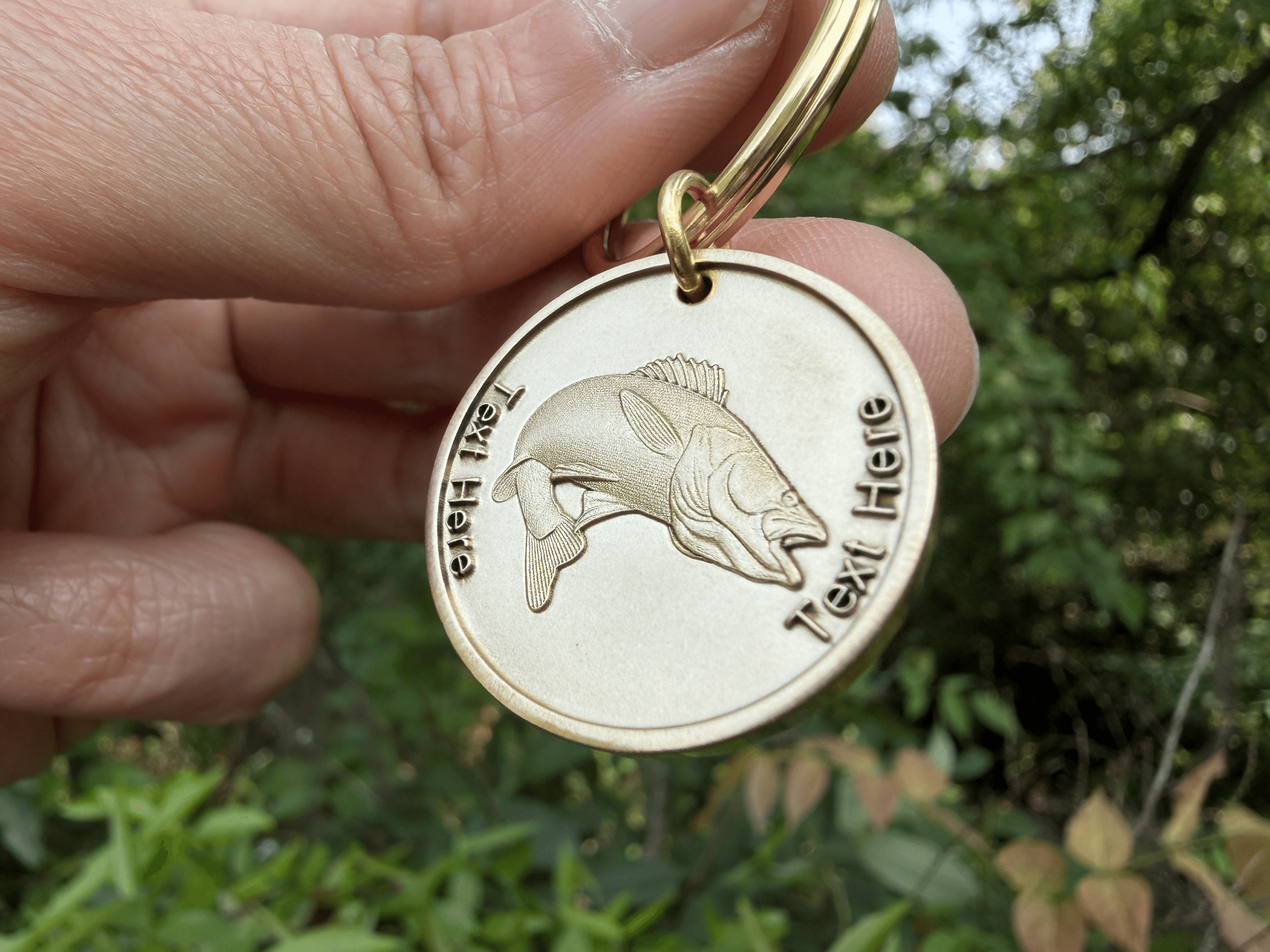 Gold keychain with engraved fish and customizable Text Here displayed against a leafy background.