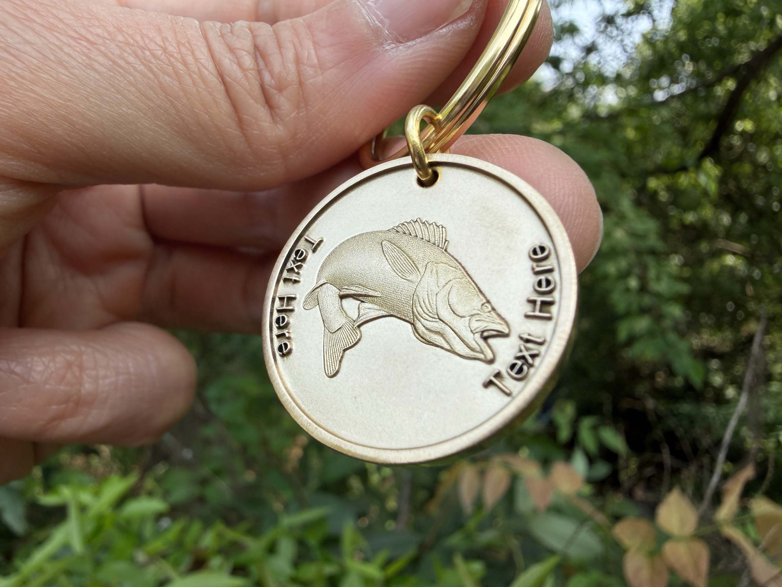 Gold keychain with engraved fish and customizable Text Here displayed against a leafy background.