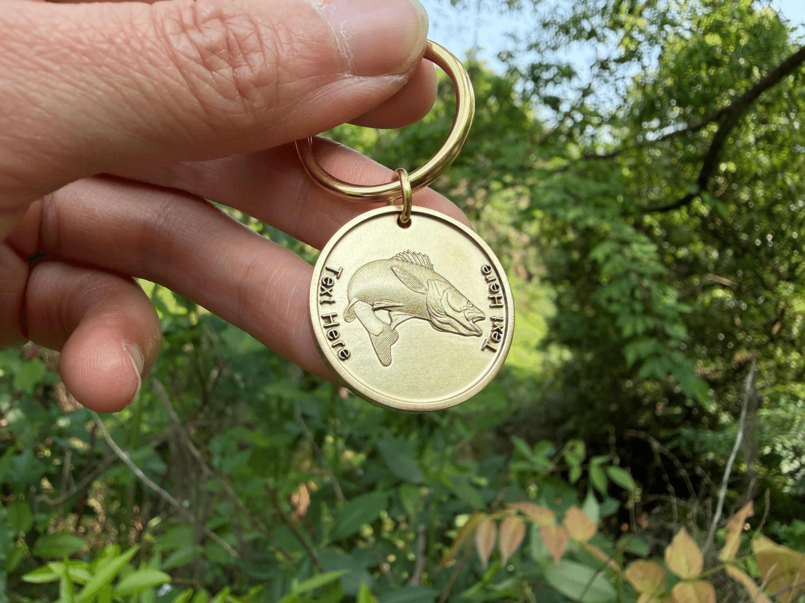 Gold keychain with engraved fish and Text Here. Hand holding it outdoors with green foliage background.