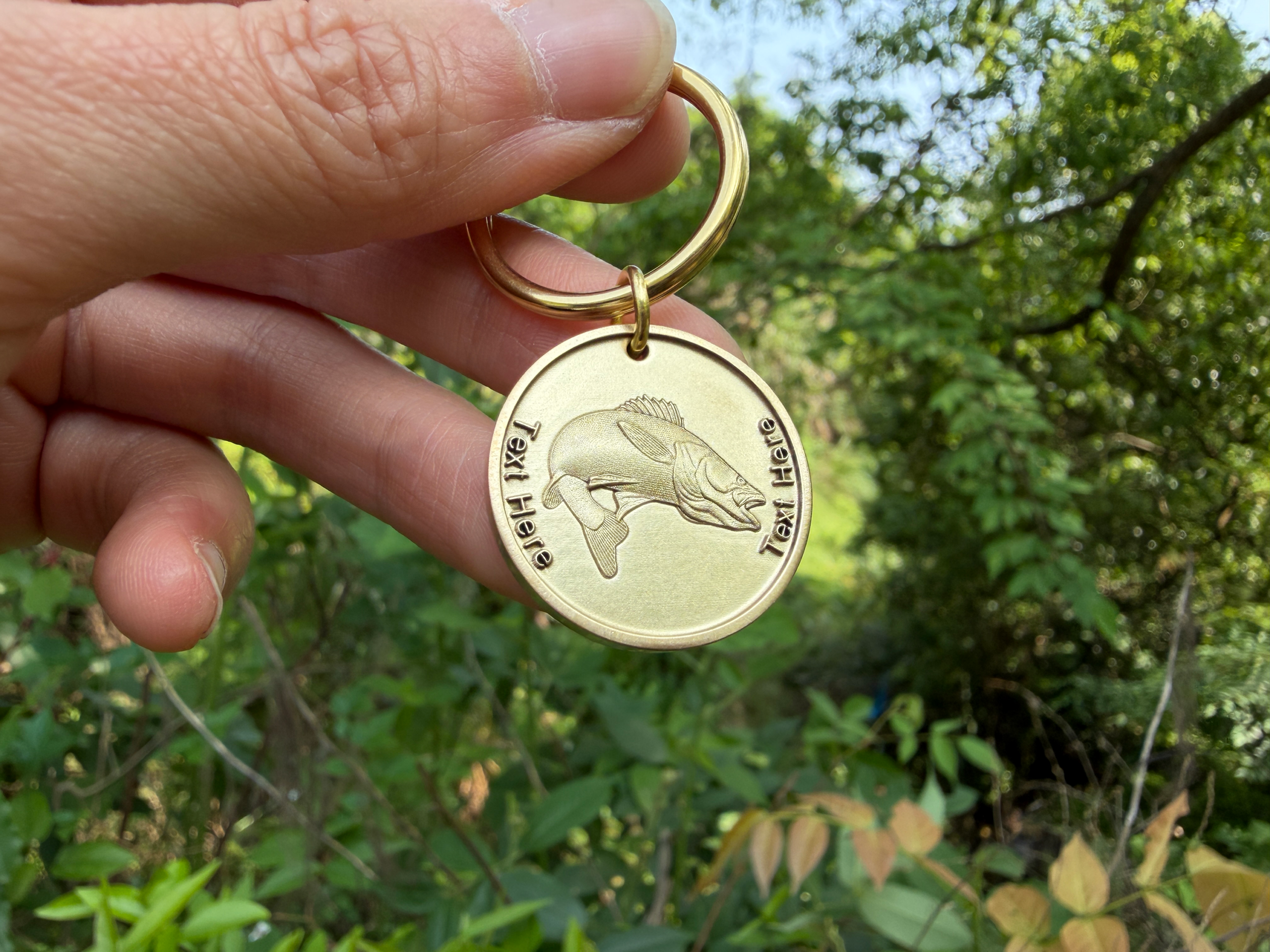 Gold keychain with engraved fish and Text Here. Hand holding it outdoors with green foliage background.