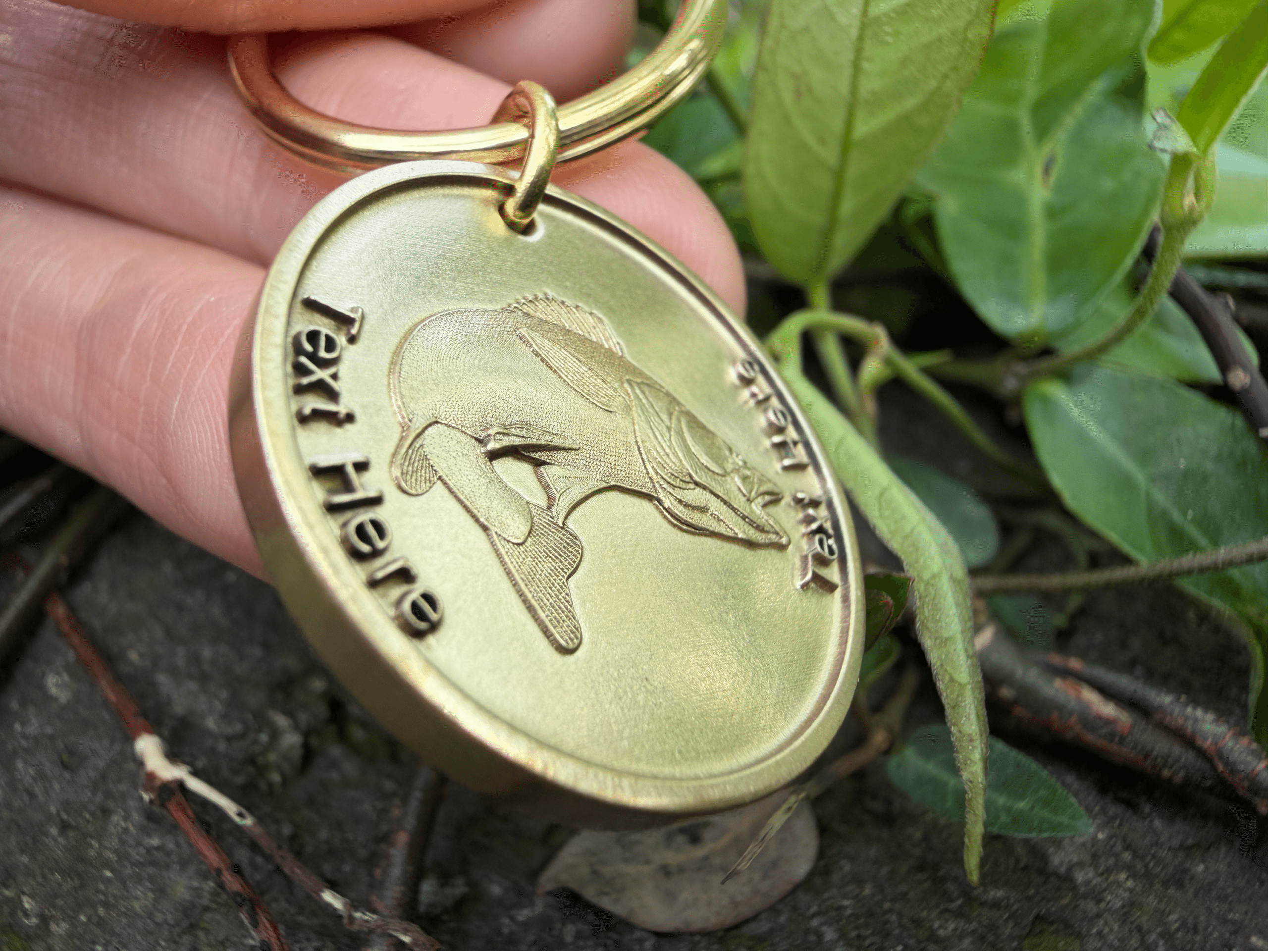 Golden keychain with bass fish engraving, held by fingers; lush green leaves in background for contrast.