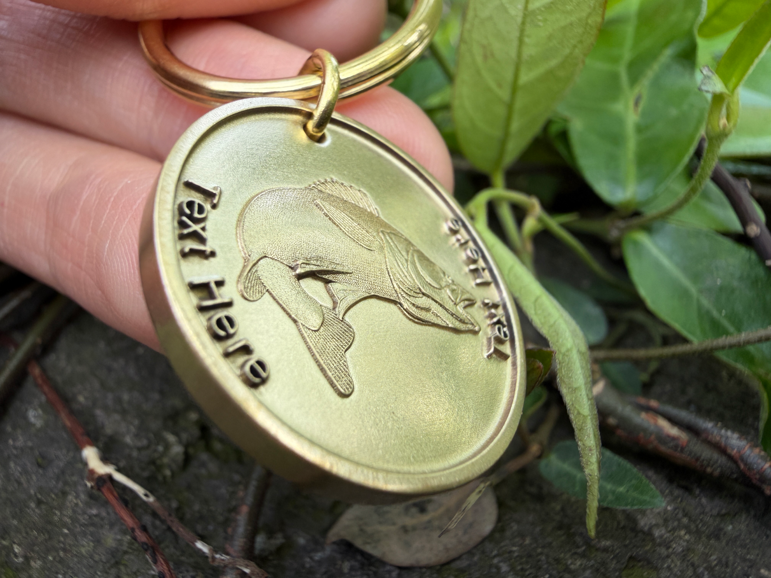 Golden keychain with bass fish engraving, held by fingers; lush green leaves in background for contrast.