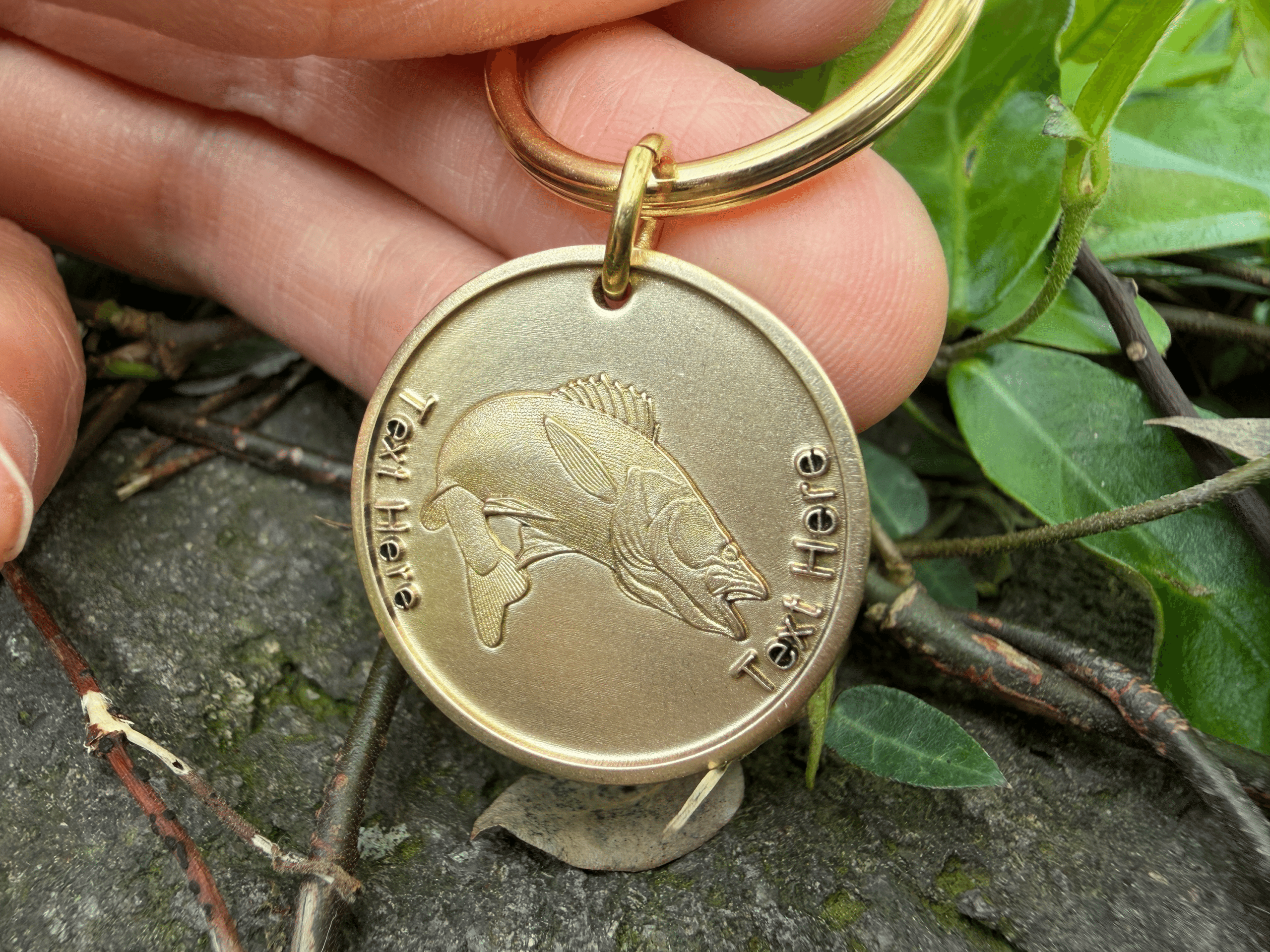 Engraved fish keychain with customizable text held by hand, surrounded by green leaves.