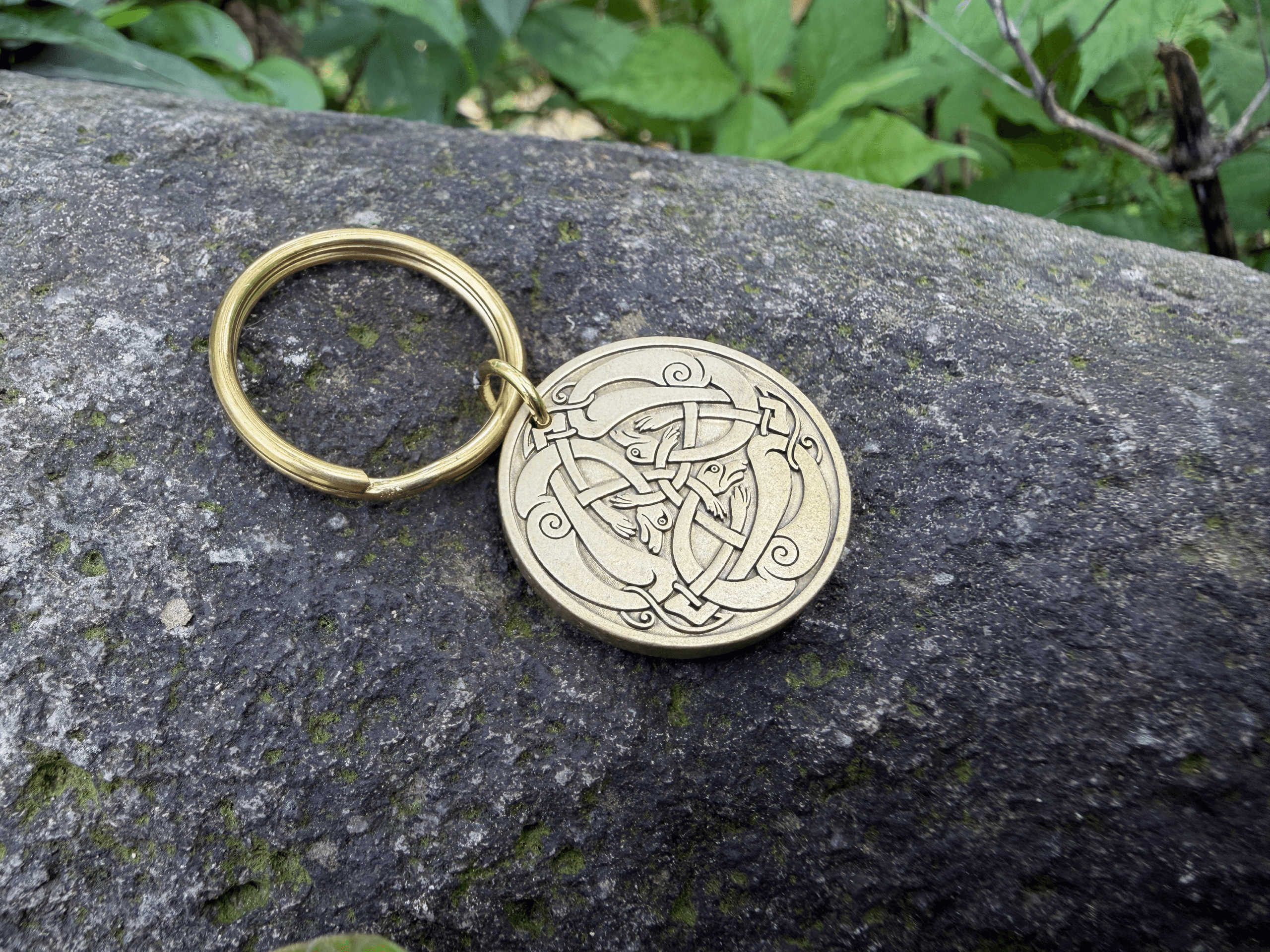 Gold keyring with intricate Odin's Horn engraving on a rock surface, surrounded by green foliage.