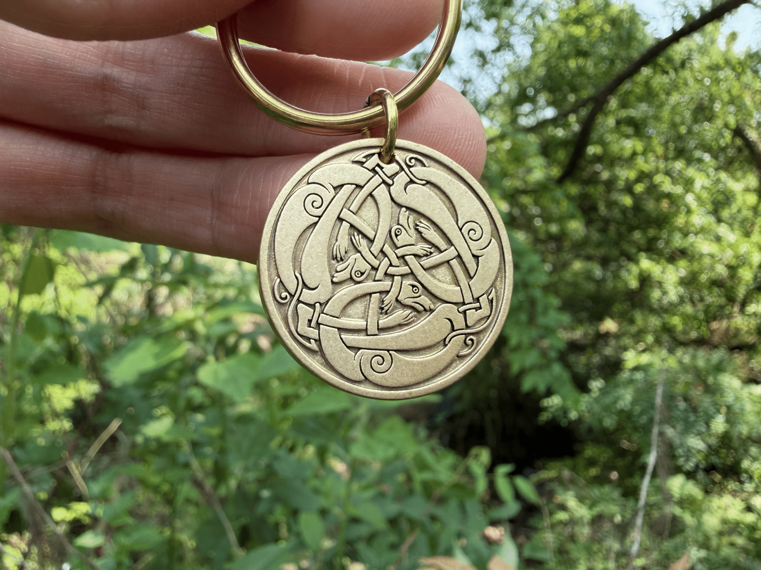 Keychain with Viking-inspired engravings of intertwined ravens, symbolizing Odin, held in hand outdoors with greenery background.