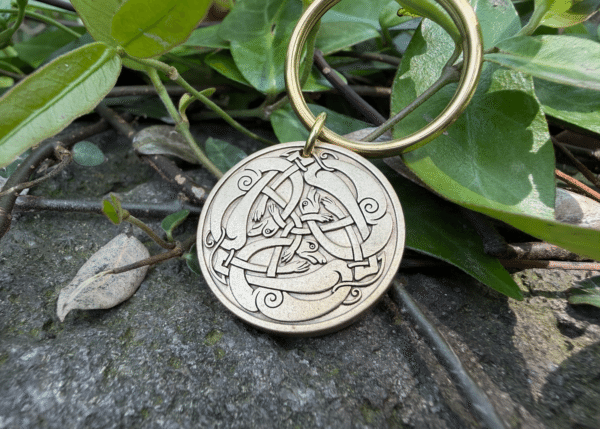 Close-up of a keychain with a brass Viking-style Jörmungandr engraving on stone with green leaves background.