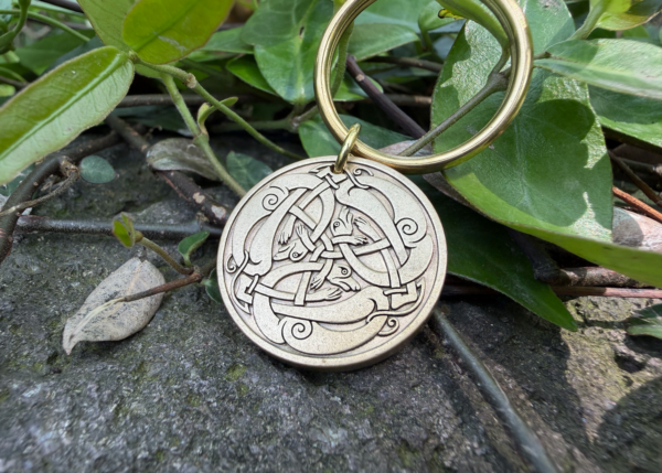 Close-up of a keychain with a brass Viking-style Jörmungandr engraving on stone with green leaves background.