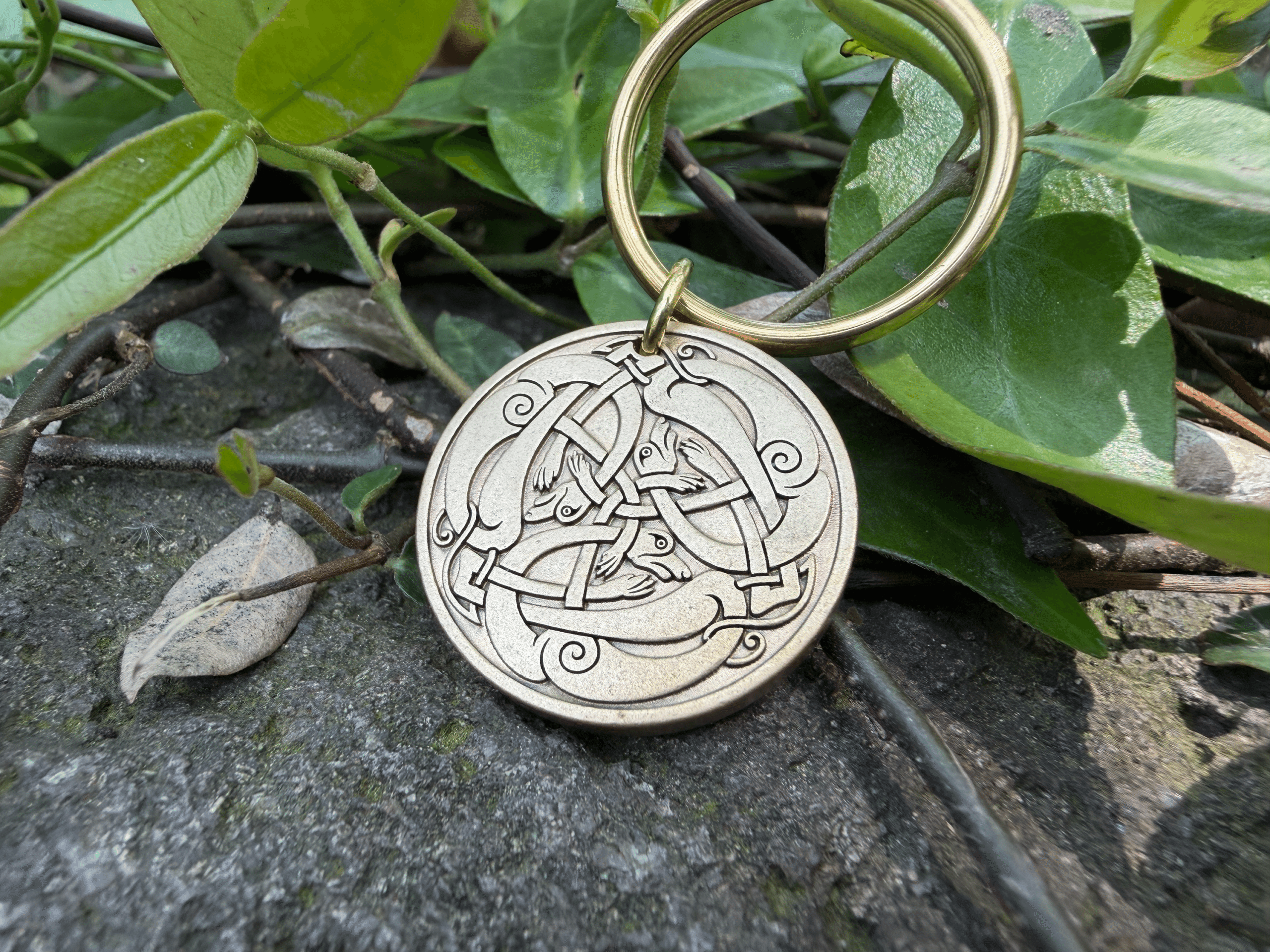 Close-up of a keychain with a brass Viking-style Jörmungandr engraving on stone with green leaves background.