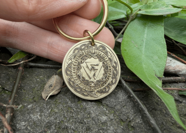 Gold keychain featuring intricate Norse runes and Viking symbols, held by hand over a natural background with leaves.