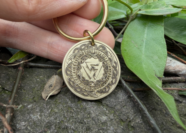 Gold keychain featuring intricate Norse runes and Viking symbols, held by hand over a natural background with leaves.