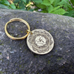 Golden keychain with intricate Norse design engraved on a coin, resting on a mossy rock in natural surroundings.