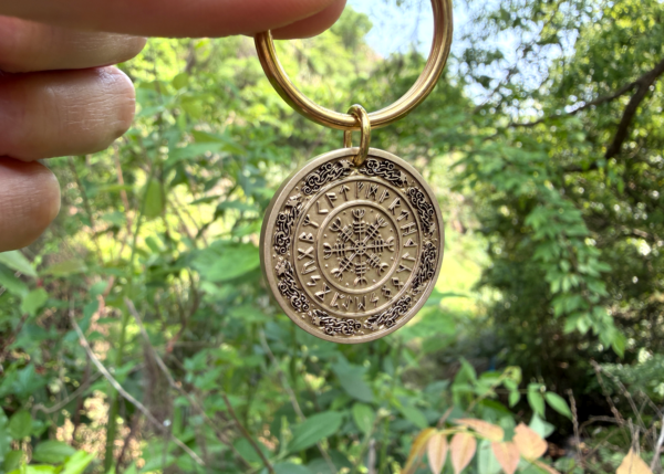 Round keychain with Vegvísir engraving held against a lush green forest background.