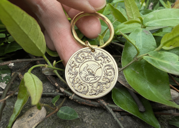 Intricate circular keychain with ocean wave engraving, surrounded by animal designs, placed on green leaves.