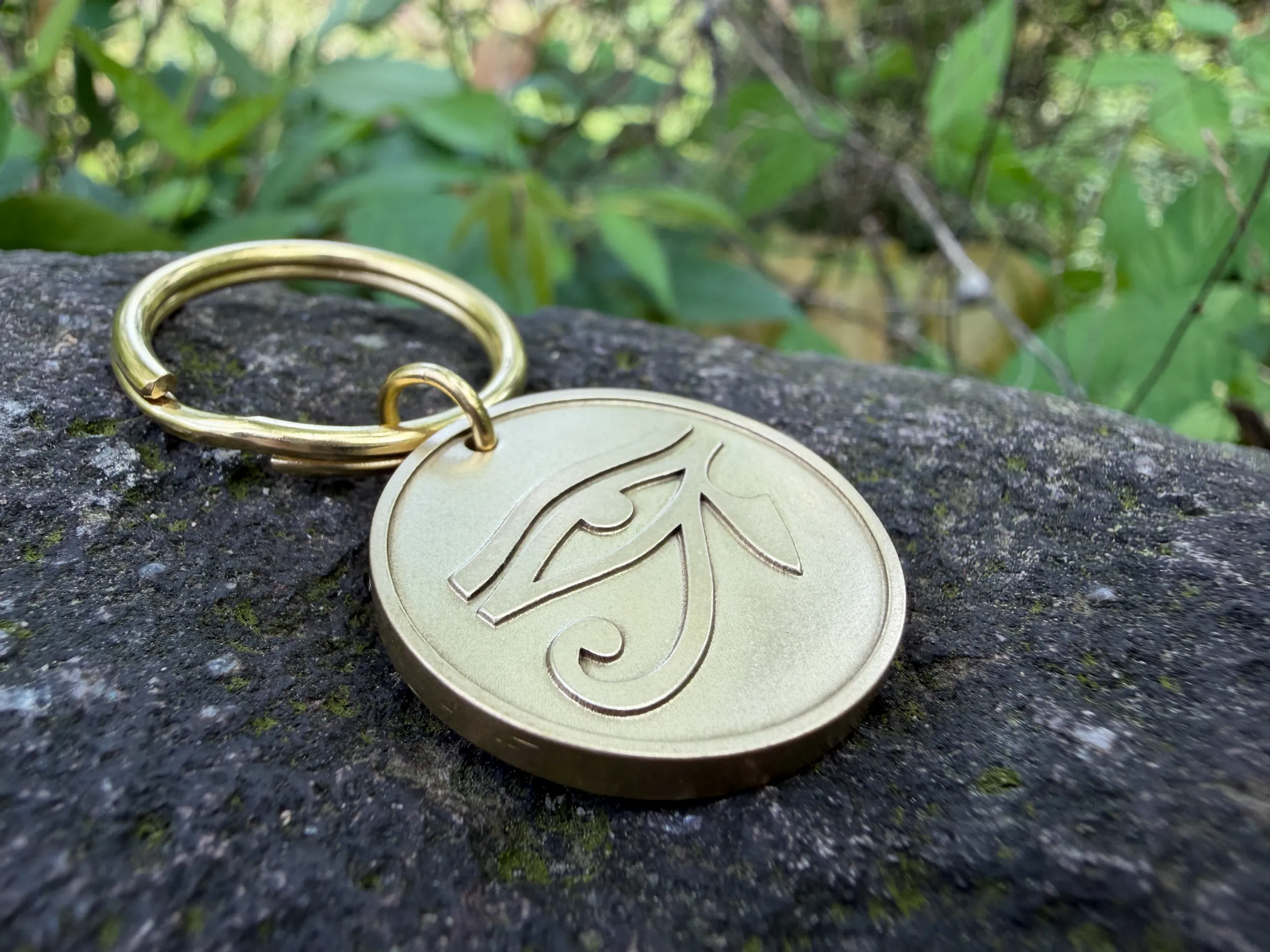 Gold keychain on stone with an engraved Eye of Horus symbol, surrounded by greenery.