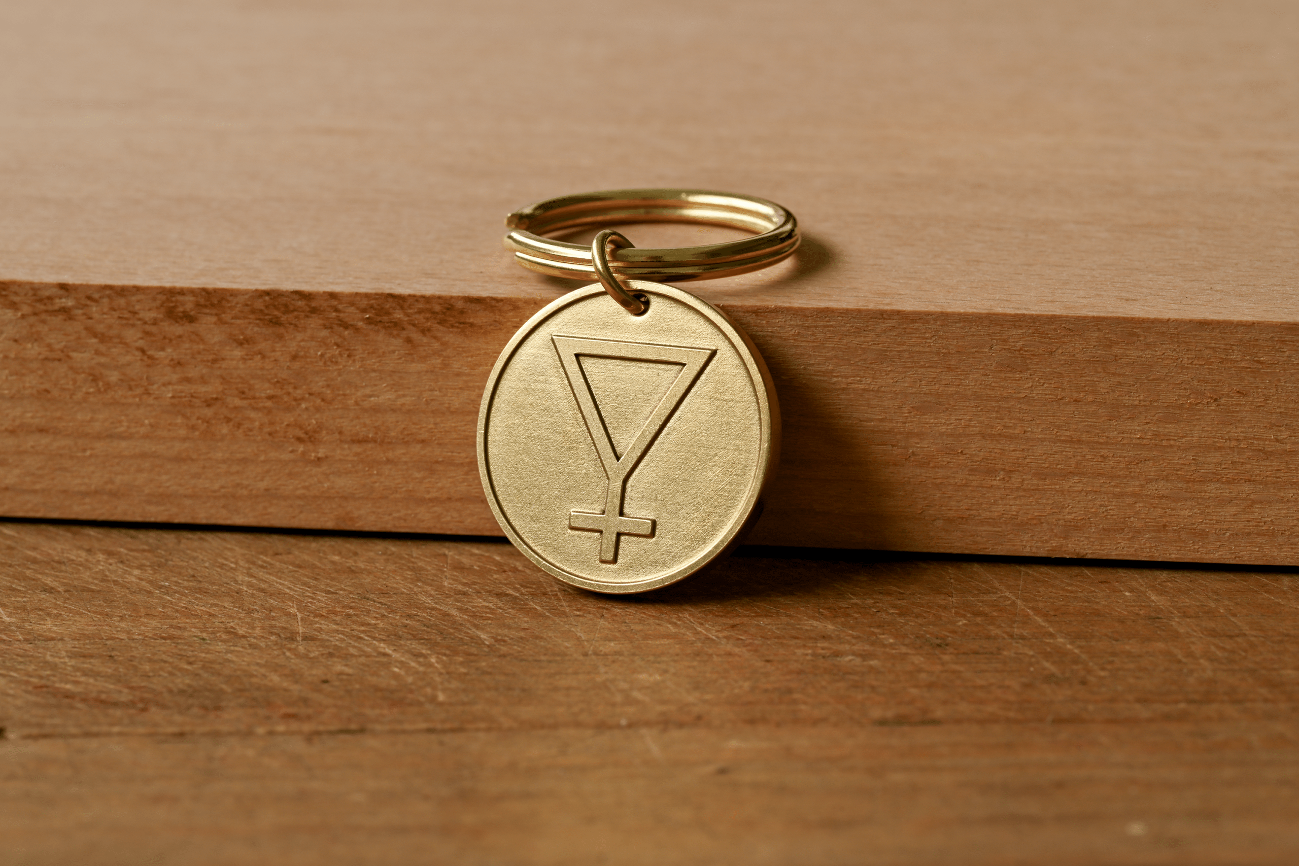 A circular brass keychain tag engraved with an inverted triangle with a cross below, resting on a wooden surface