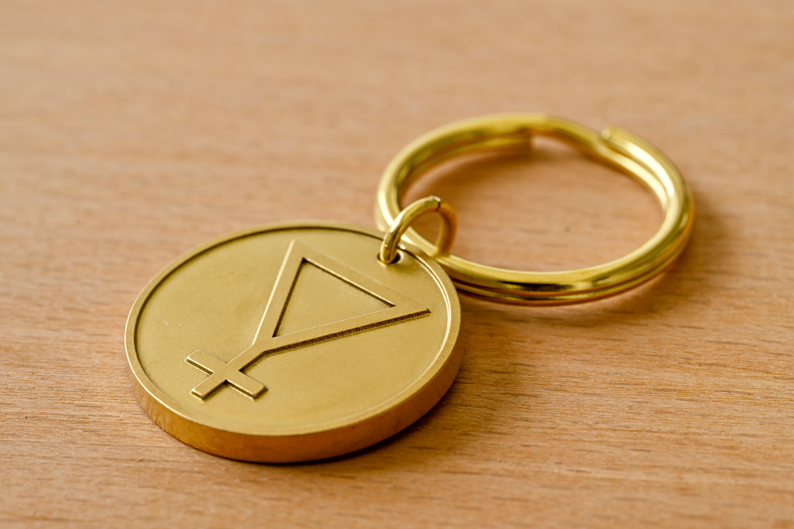 Gold metal keychain pendant engraved with an alchemical symbol, resting on a wooden surface