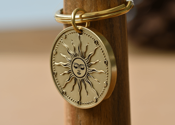 Round brass keychain pendant featuring an engraved celestial sun face with wavy rays and a moon phase border