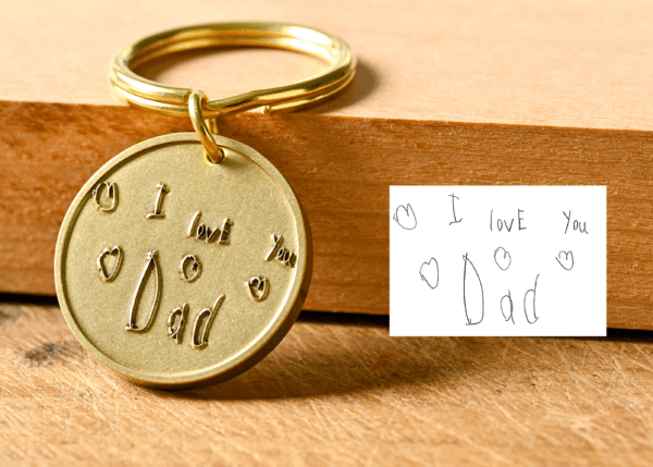 Gold keychain engraved with handwritten text reading I love You Dad alongside the original note on paper