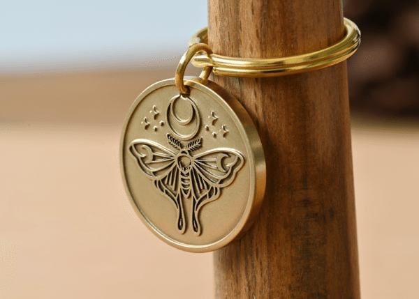 Round brass keychain with an engraved moth design under a crescent moon and small stars