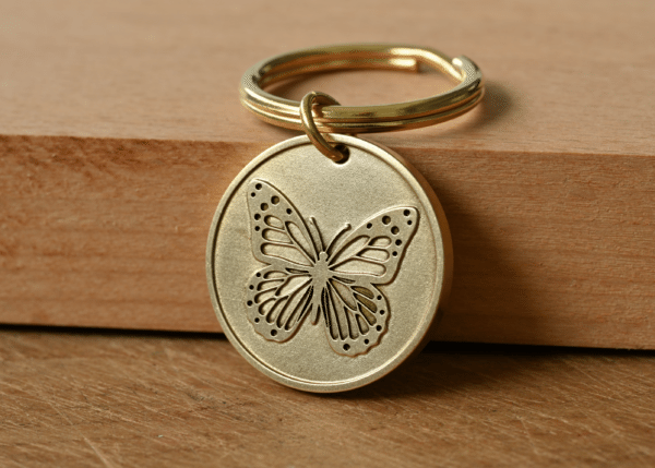 Round brass keychain tag featuring a detailed engraved butterfly, resting on a wooden block