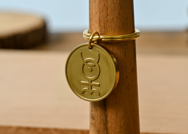 Gold brass keychain tag engraved with an alchemical symbol, displayed on a wooden post
