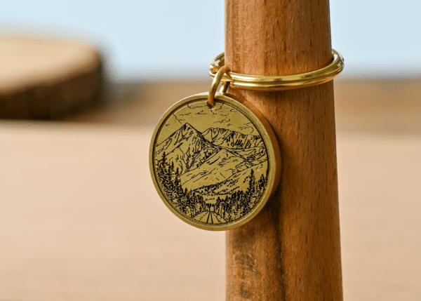 Engraved brass keychain featuring a mountain landscape with pine trees and a road