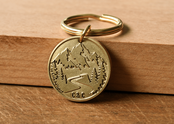 Round brass keychain engraved with mountains, pine trees, a winding path, and the initials C & C