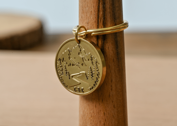 Gold circular keychain tag engraved with a mountain landscape, pine trees, winding road, and initials C & C