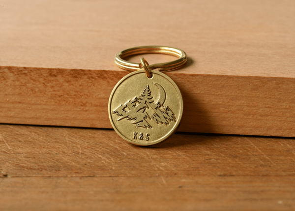 Round brass keychain engraved with mountain peaks, pine trees, a crescent moon, and the initials K&S
