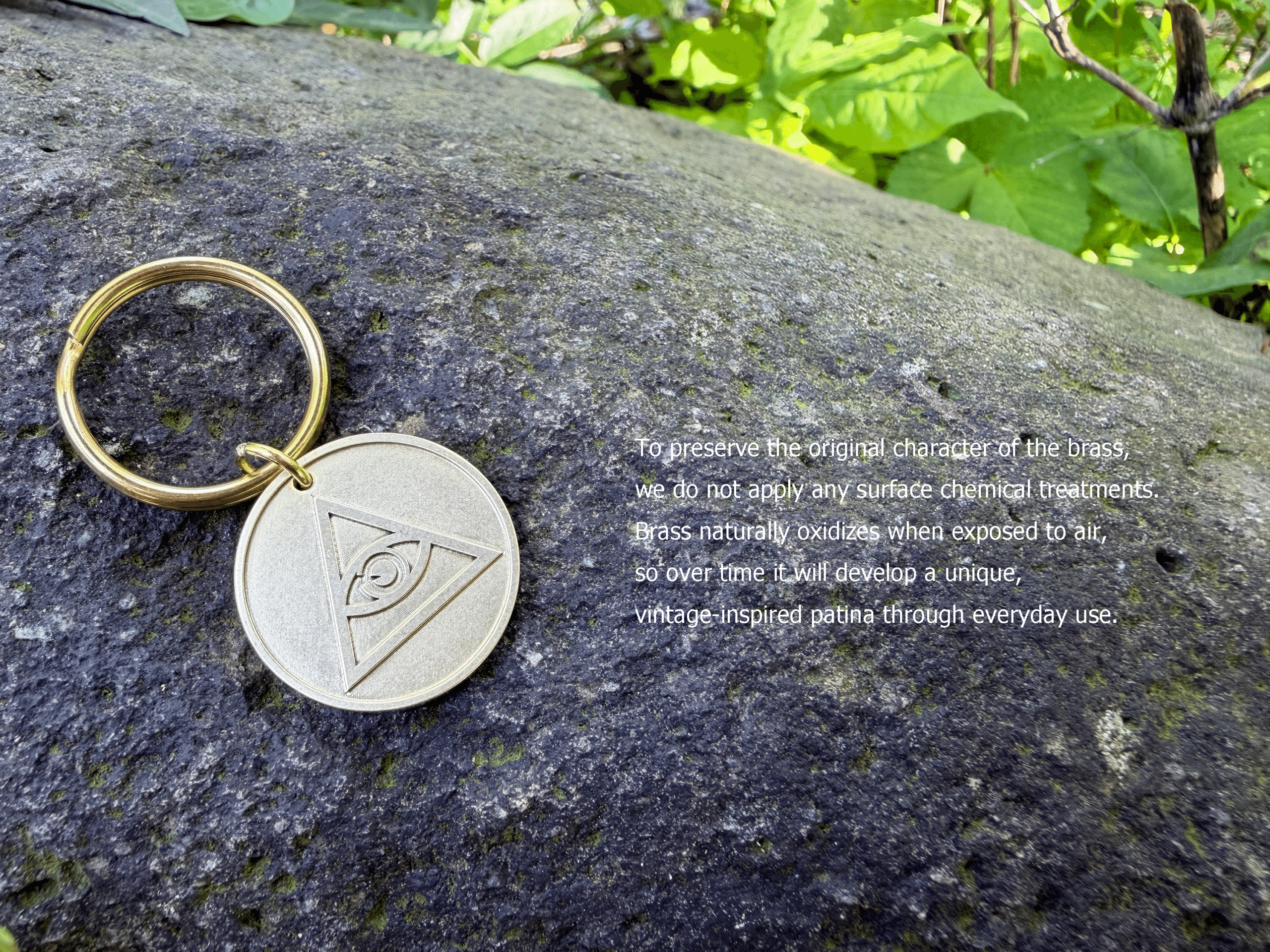 Brass Eye of Providence keychain resting on a rock. Text: To preserve the original character of the brass, we do not apply any surface chemical treatments. Brass naturally oxidizes when exposed to air, so over time it will develop a unique, vintage-inspired patina through everyday use