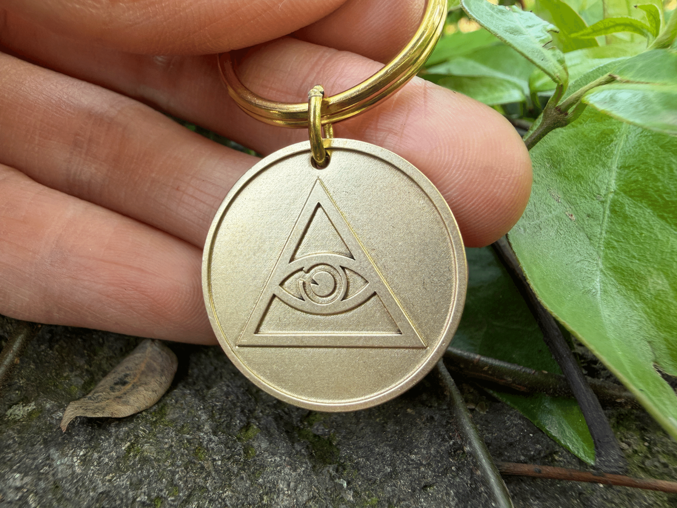 Hand holding a round brass keychain medallion engraved with an Eye of Providence symbol
