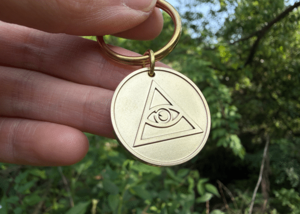 Hand holding a brass keychain featuring a circular medallion engraved with the Eye of Providence symbol