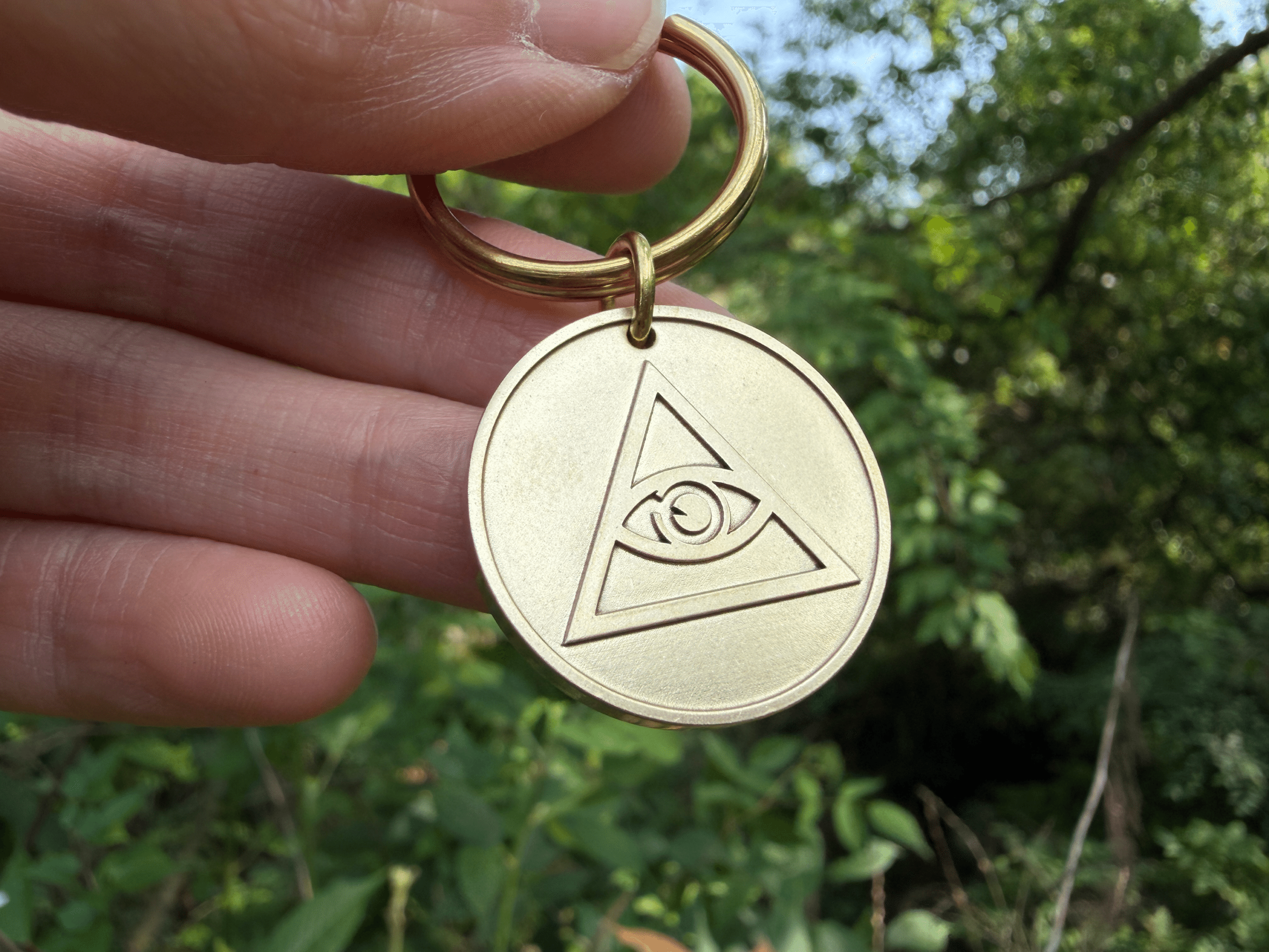Hand holding a brass keychain featuring a circular medallion engraved with the Eye of Providence symbol
