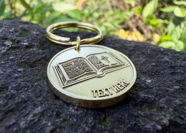 Round brass keychain engraved with an open Bible featuring a cross and placeholder text reading TEXT HERE