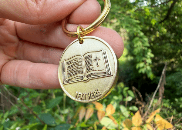 Gold circular keychain medallion engraved with an open Bible, a cross, and placeholder text reading Text Here