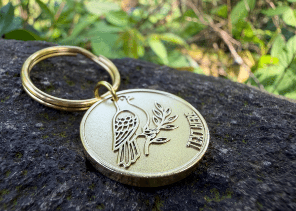 Circular brass keychain tag engraved with a bird perched on a leafy branch alongside placeholder text