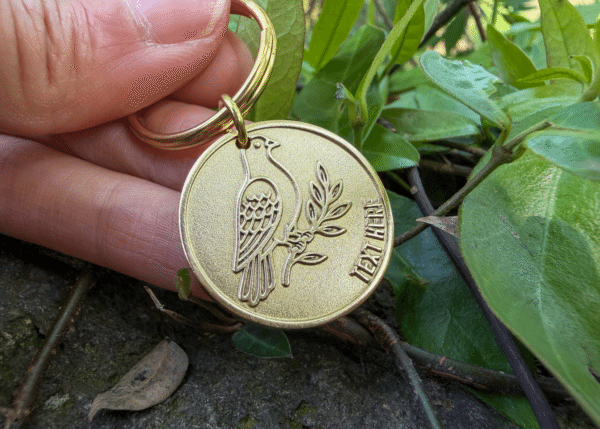 Round brass keychain engraved with a bird on a branch and the text TEXT HERE
