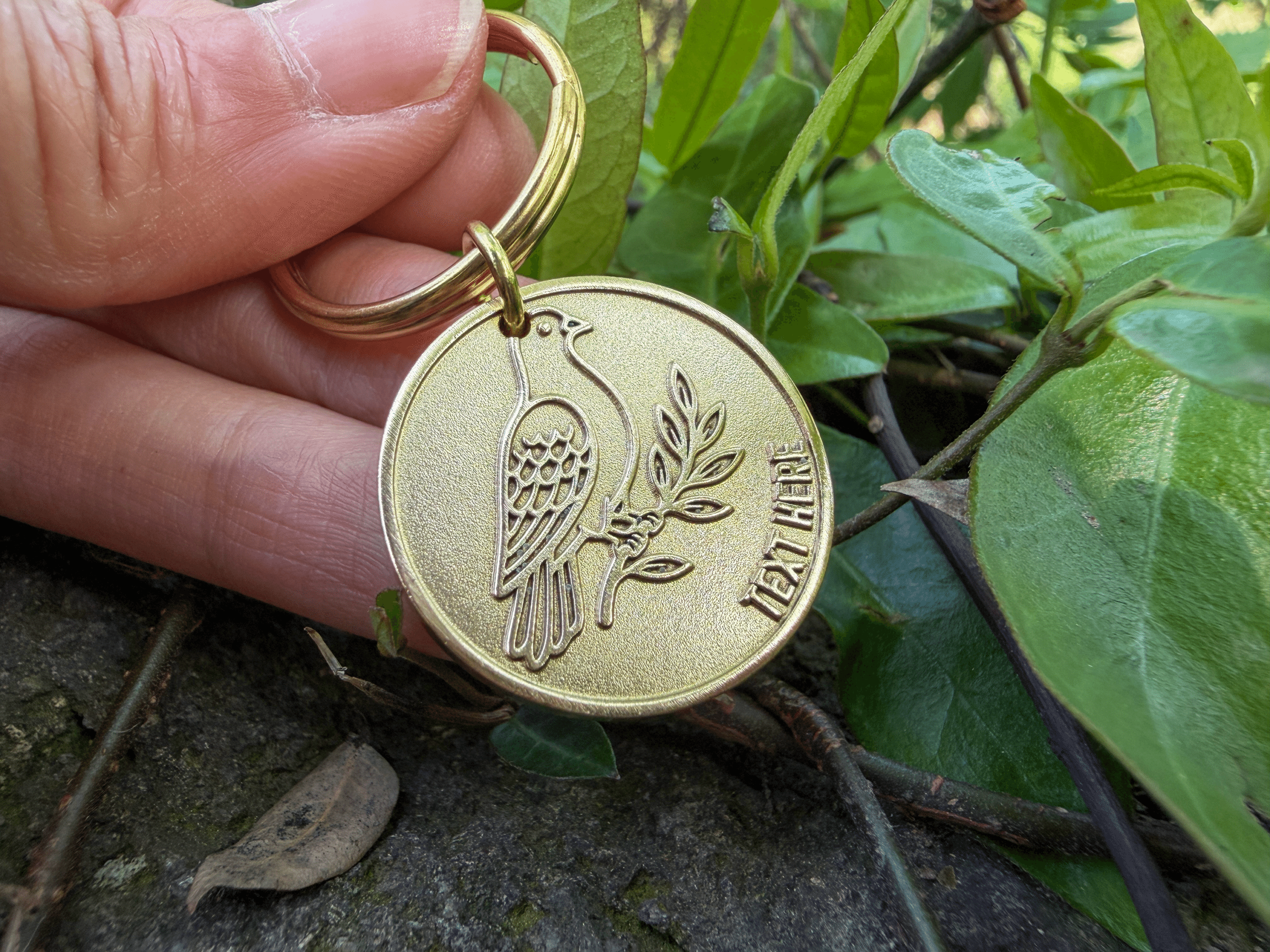 Round brass keychain engraved with a bird on a branch and the text TEXT HERE