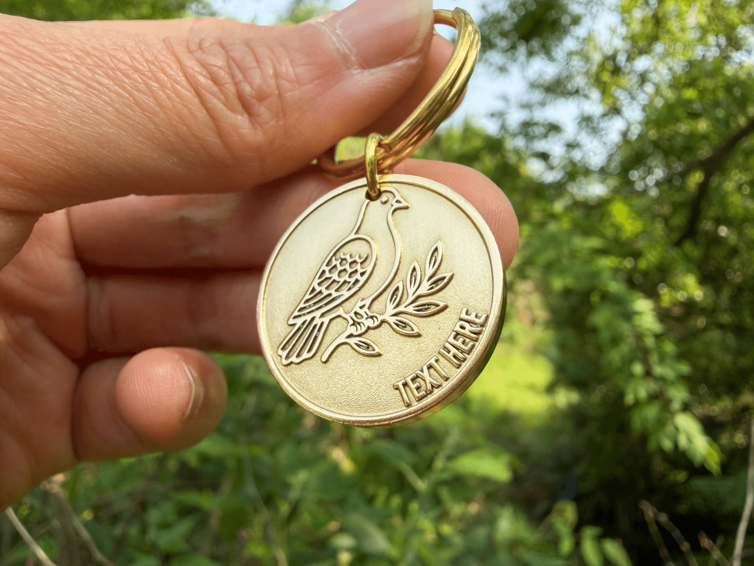 Hand holding a round gold keychain with an engraved bird on a branch and text reading TEXT HERE