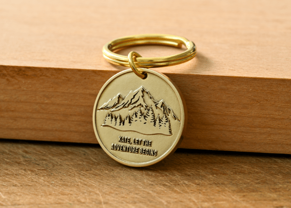 Round brass keychain with an engraved mountain and forest scene, featuring the text Kate, Let The Adventure Begins