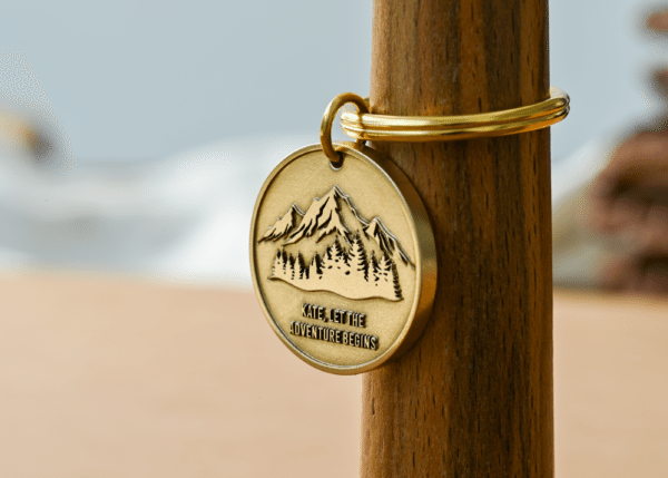 Engraved brass keychain with mountains, trees, and text reading Kate, let the adventure begins