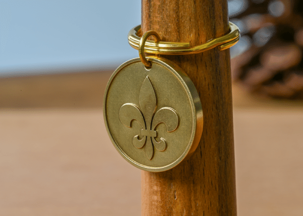 Brass circular keychain pendant with an embossed fleur-de-lis symbol hanging on a wooden post