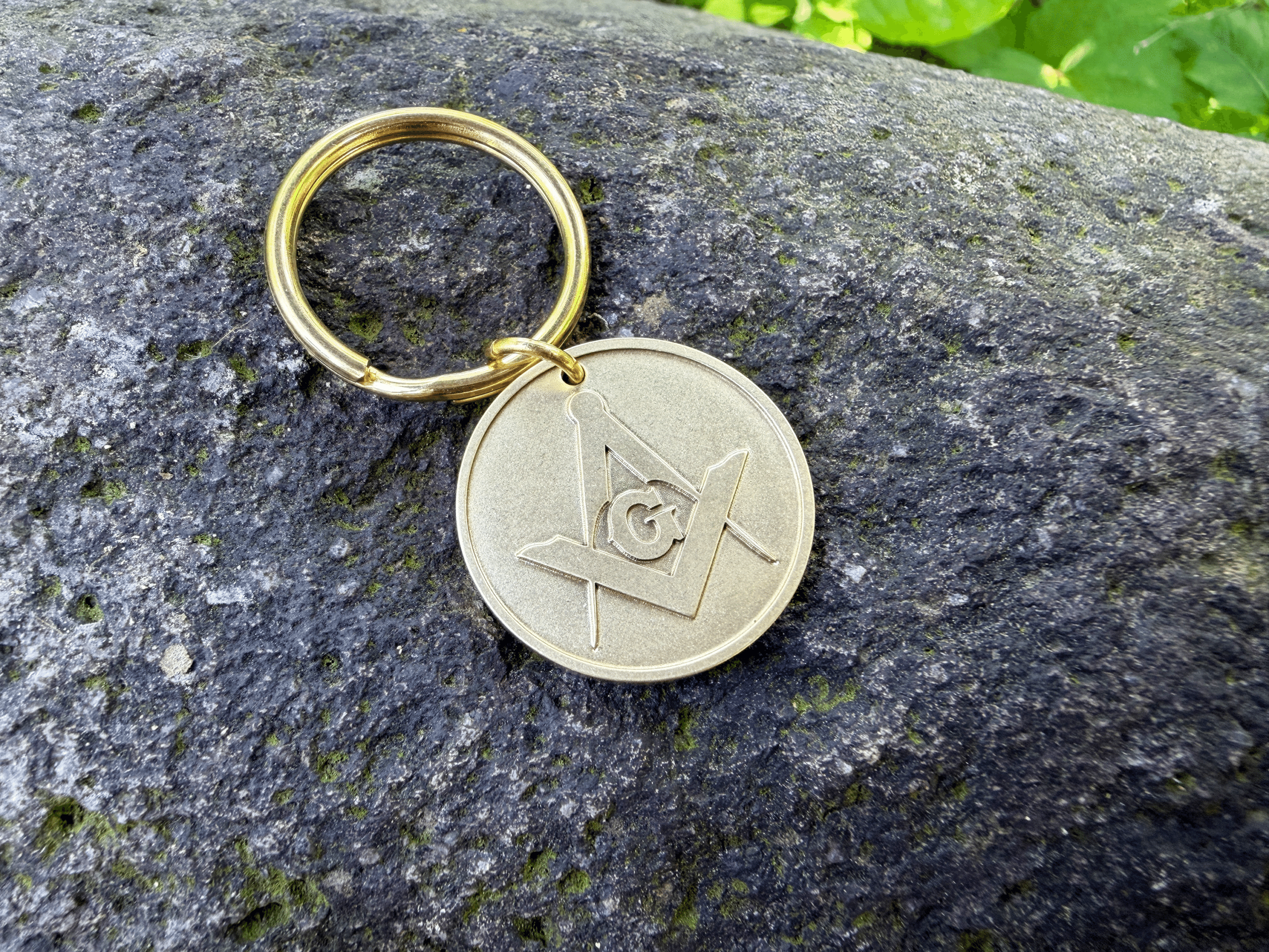 Gold keychain medallion engraved with the Masonic Square and Compasses symbol resting on a stone surface