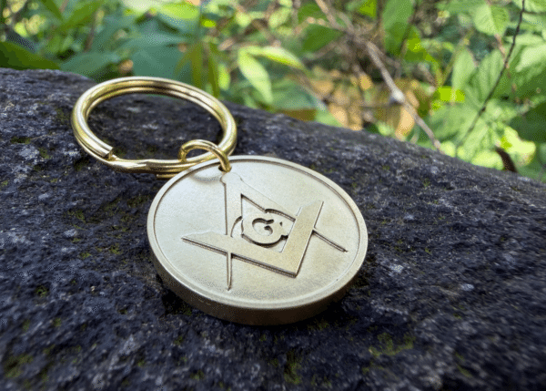 Brass keychain with the Masonic Square and Compasses symbol resting on a rock