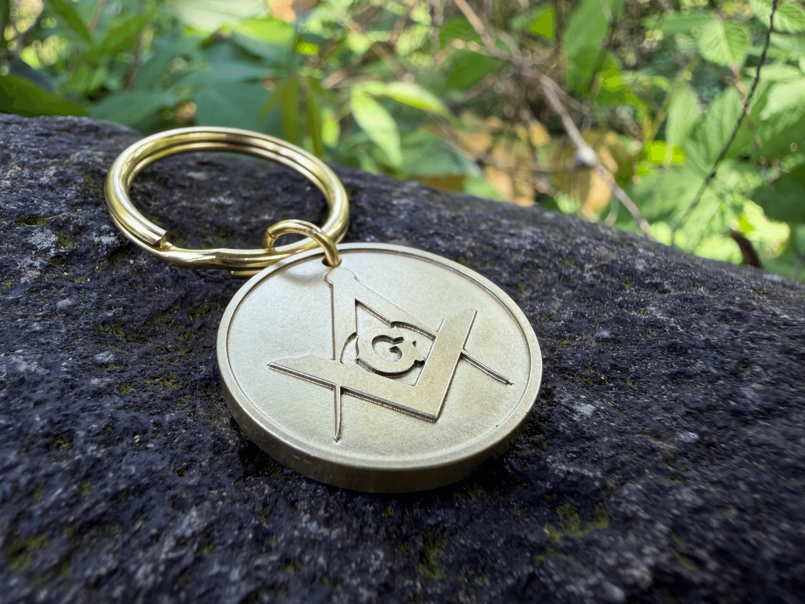 Brass keychain with the Masonic Square and Compasses symbol resting on a rock