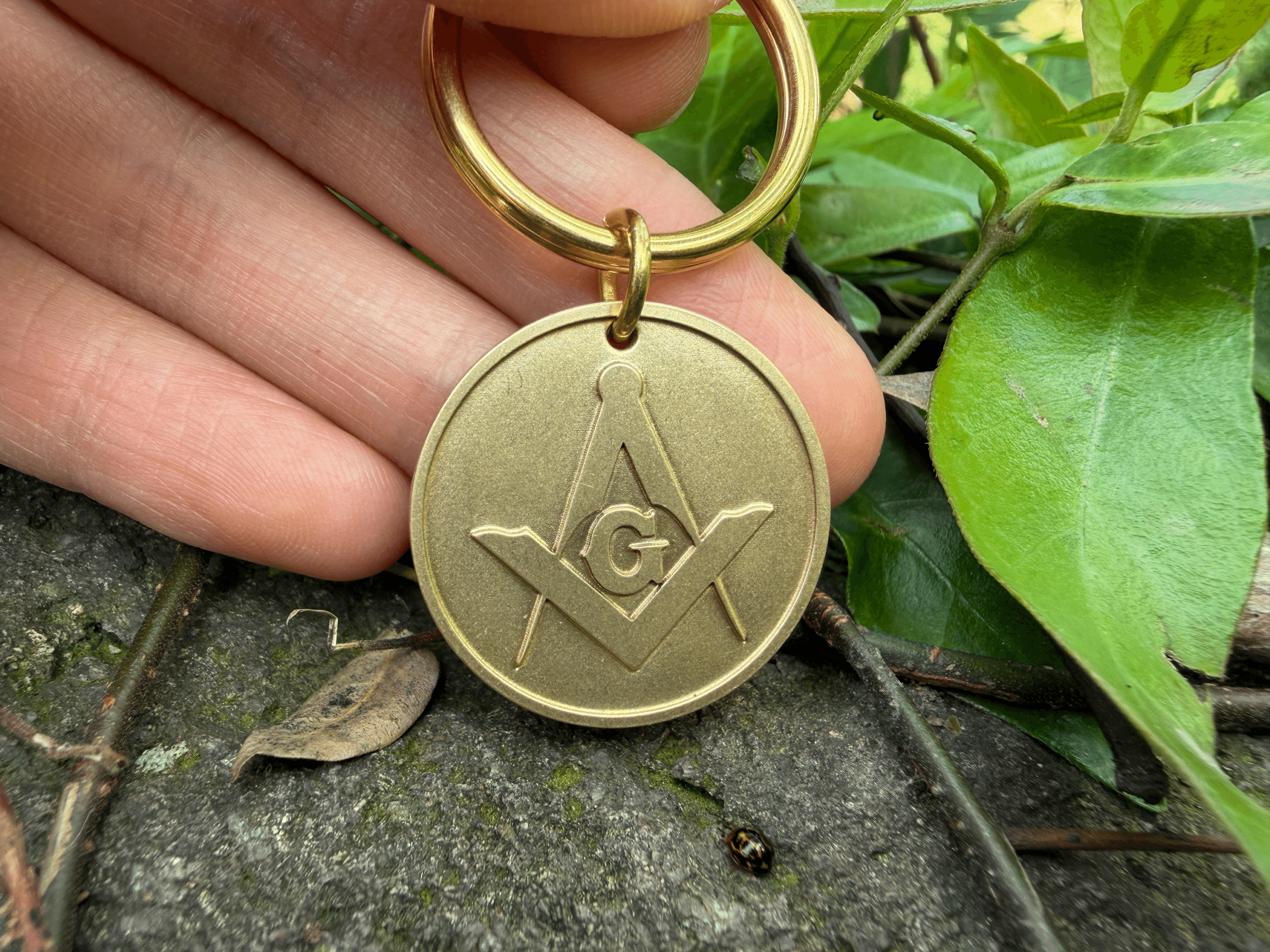 Hand holding a brass circular keychain medallion engraved with the Masonic Square and Compasses symbol with the letter G